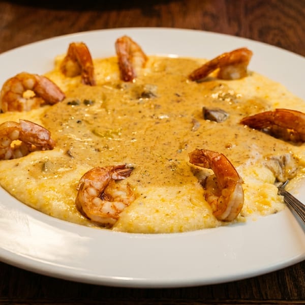 Creamy shrimp and grits garnished with sautéed shrimp, served on a white plate atop a wooden restaurant table.