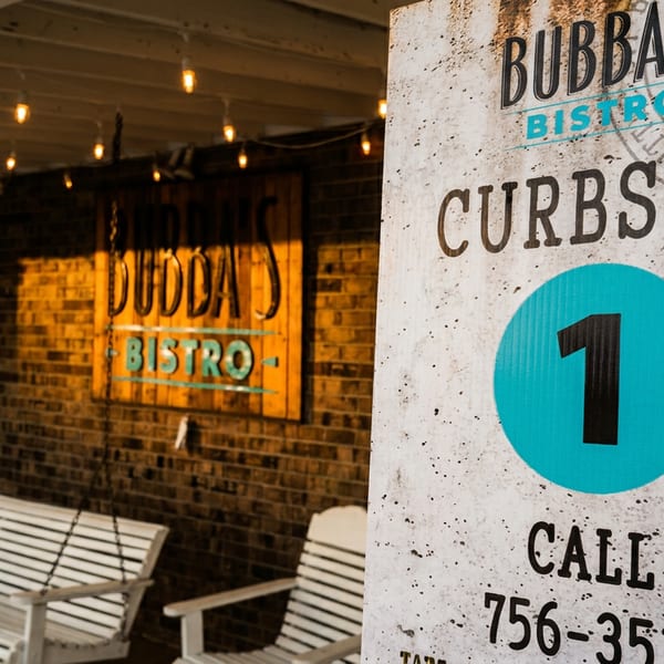 Bubba’s Bistro curbside pickup sign outside a warmly lit brick restaurant with white swing chairs.