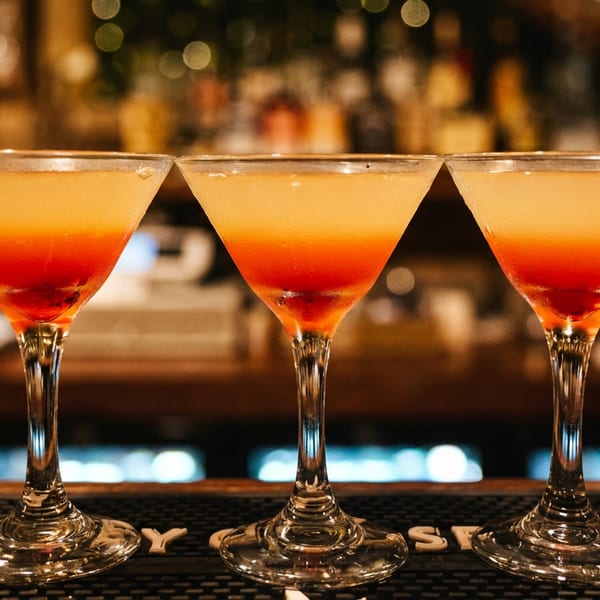 Three orange-red cocktails in martini glasses sit on a bar mat against a warmly lit, blurred bar background.