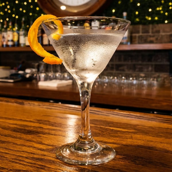 Chilled martini garnished with an orange peel sits on a polished wooden bar in a warmly lit restaurant.