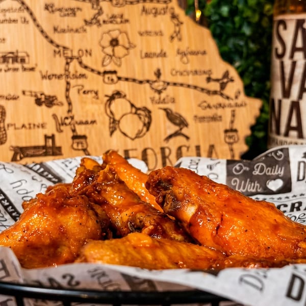 Glazed orange chicken wings piled in a basket lined with deli paper, with a rustic map and beer bottle in the background.