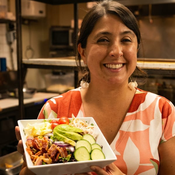 Smiling woman in a restaurant kitchen holds a bowl featuring bacon, avocado, cucumber, tomatoes, greens, and shredded vegetables.
