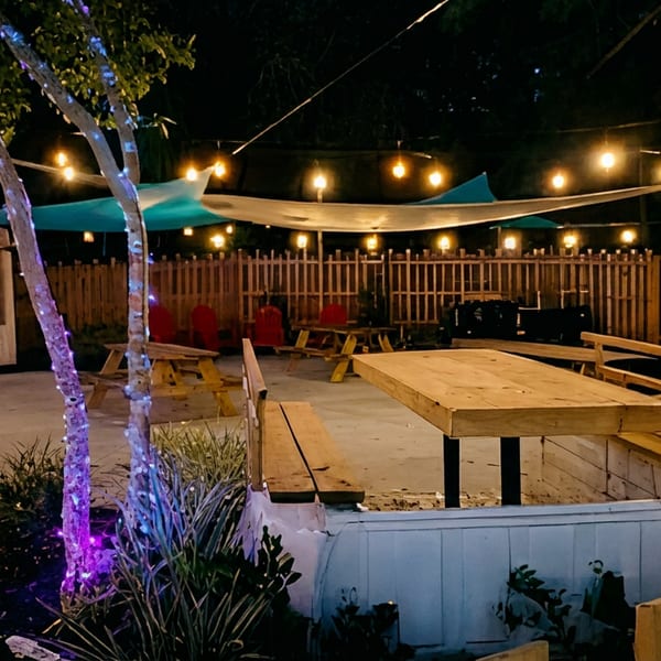 Outdoor restaurant patio at night with picnic tables, colorful shade sails, string lights, and illuminated trees.