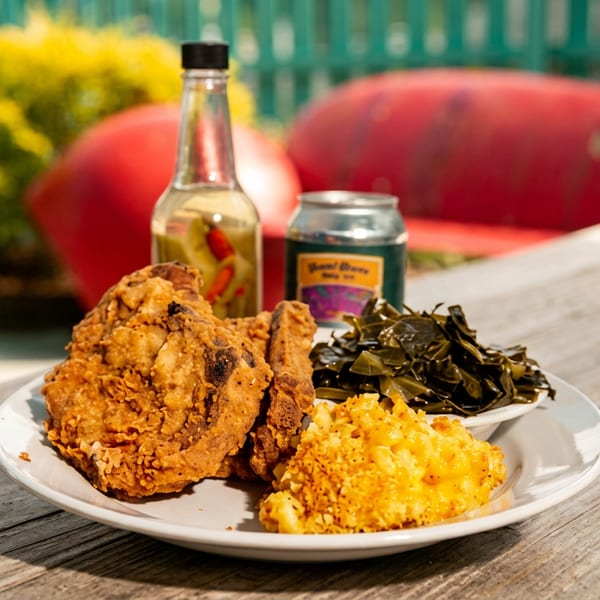 Plate of crispy fried chicken, macaroni and cheese, and greens on a wooden outdoor table with hot sauce bottles.