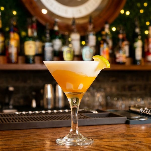 Orange cocktail in a martini glass garnished with a lime wedge, displayed on a warmly lit bar with bottles and mixing tools.