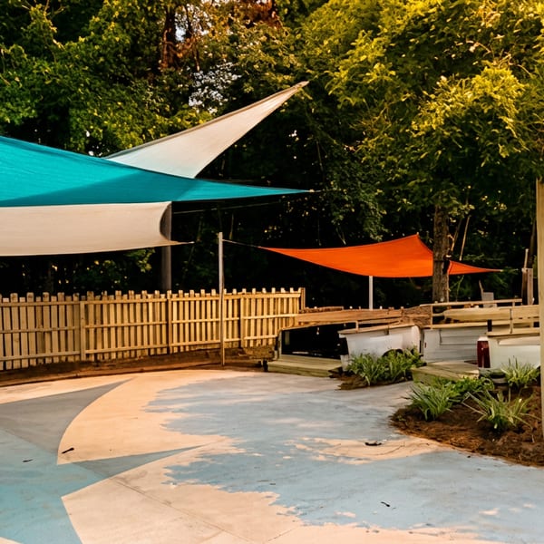 Outdoor restaurant seating area with colorful shade sails, wooden fencing, raised planters, and trees.