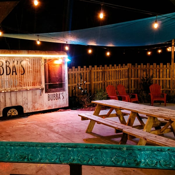 Bubba’s food truck sits in a warmly lit outdoor dining area with picnic tables, red chairs, string lights, and a wooden fence.
