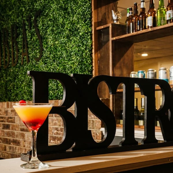 Colorful cocktail garnished with a cherry sits before a large black BISTRO sign, with bottles displayed on wooden shelves.