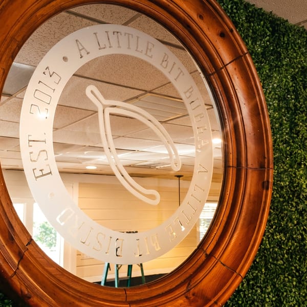 Ornate wooden oval mirror with a circular restaurant logo, surrounded by greenery and reflecting a warmly lit dining interior.
