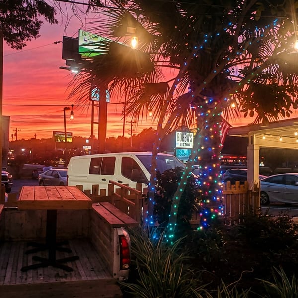 Outdoor restaurant patio at sunset with picnic tables, string-lit palm trees, parked vehicles, and a visible Bubba’s Bistro sign.
