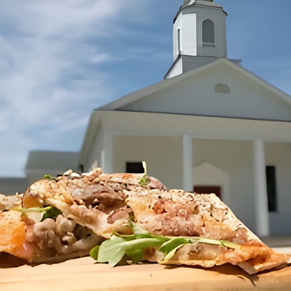 Southern-style sandwich with meat and greens on a wooden board, with a white church in the background.