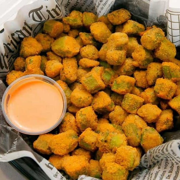 A basket of golden-fried okra bites with a side of creamy dipping sauce, lined with newspaper.