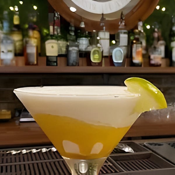 A yellow and white layered cocktail with a lime wedge, on a bar with liquor bottles.