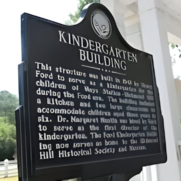 A black historical marker sign for the "Kindergarten Building" with white text, against a white building.