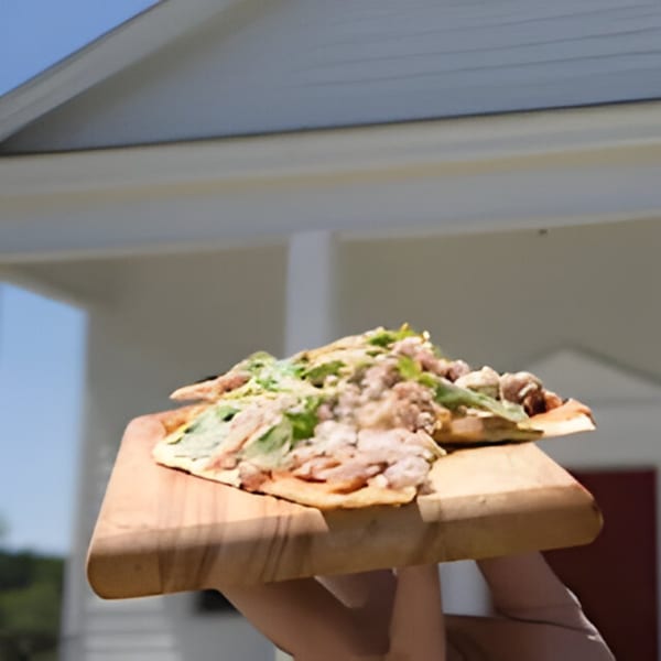 A hand holding a wooden board with a southern flatbread topped with meat and greens.