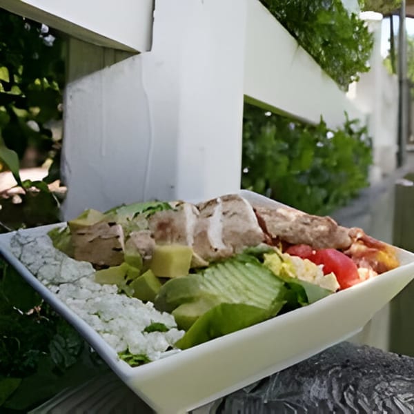A Bubba's Bistro salad with chicken, avocado, and crumbled cheese in a white bowl outdoors.
